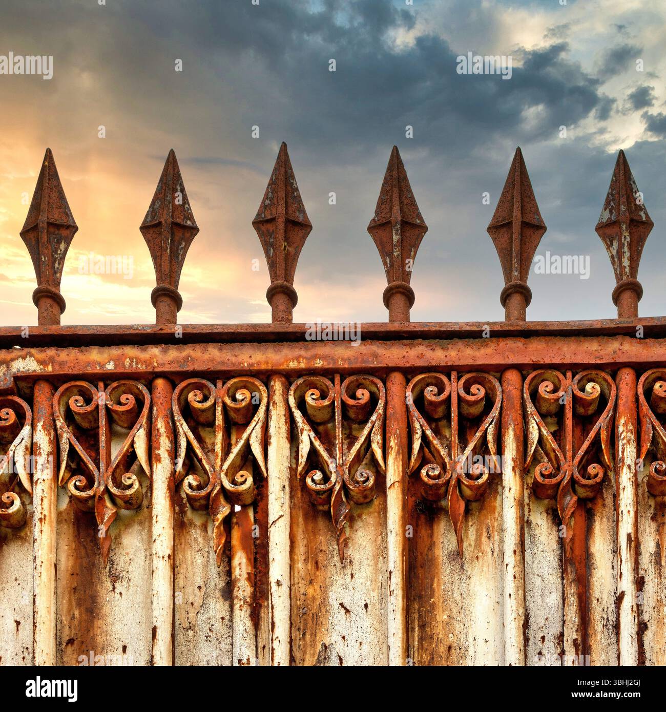 Ein rostiges, reich verziertes Tor steht hoch, mit komplizierten Designs und spitzen Spitzen. Die Kulisse zeigt einen dramatischen Himmel, der während des Sonnenuntergangs mit Wolken geschmückt ist. Stockfoto