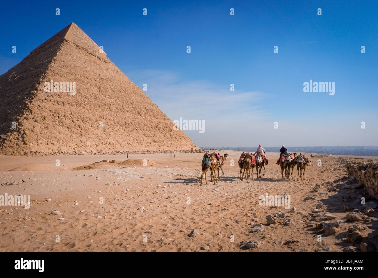 Kamelreiter durchqueren die Wüste von Gizeh mit der Pyramide von Khafre unter Einem klaren blauen Himmel in Ägypten Stockfoto