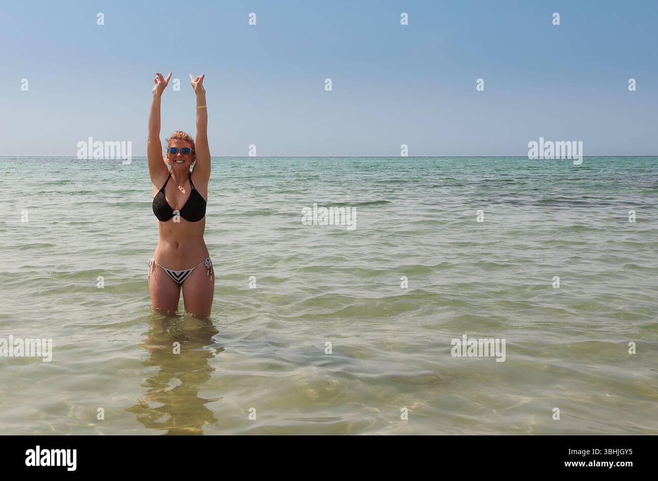 Eine kaukasische Frau in schwarz-weißem Bikini, die in klarem Meerwasser mit erhobenen Armen steht, blauer Himmel, und den Sommer am Strand in Cornwall, Großbritannien genießt Stockfoto