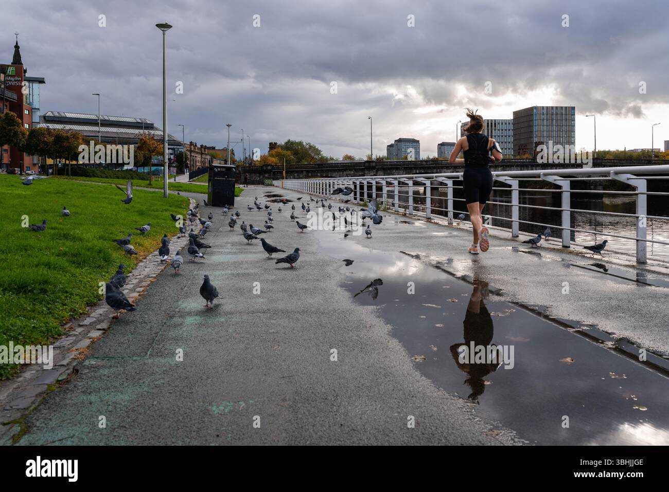 Ein Läufer bewegt sich an einem bewölkten Tag entlang der Glasgower River Clyde Promenade, vorbei an Pfützen und Tauben, mit Stadtgebäuden im Hintergrund. Stockfoto