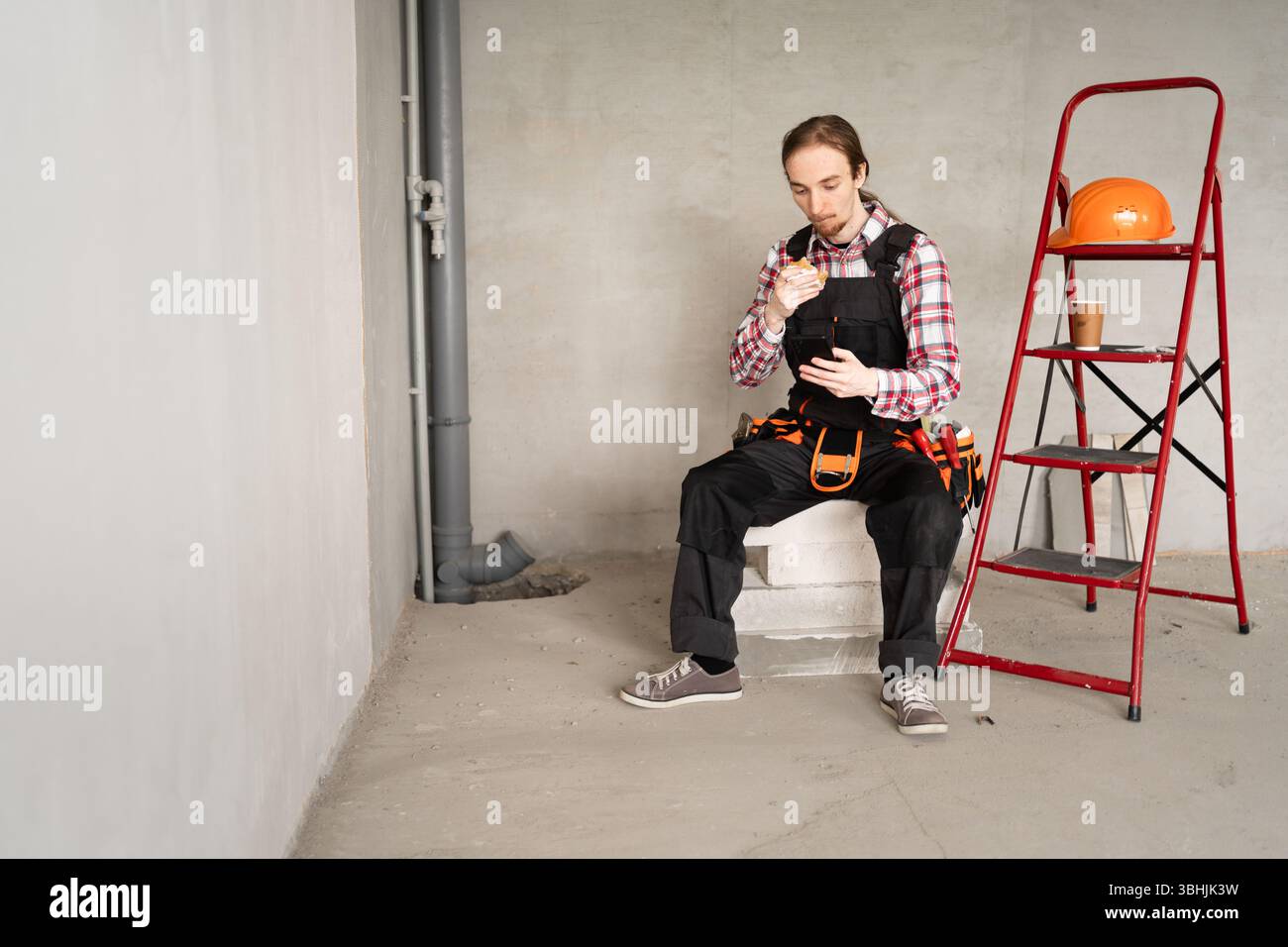 Junger Baumeister isst Burger mit Smartphone drinnen Stockfoto
