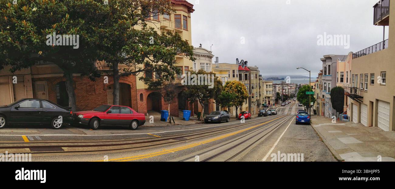 Breites Panoramabild mit einer bergab städtischen Straße mit Blick auf den Hafen, Bäumen und geparkten Autos. Stockfoto