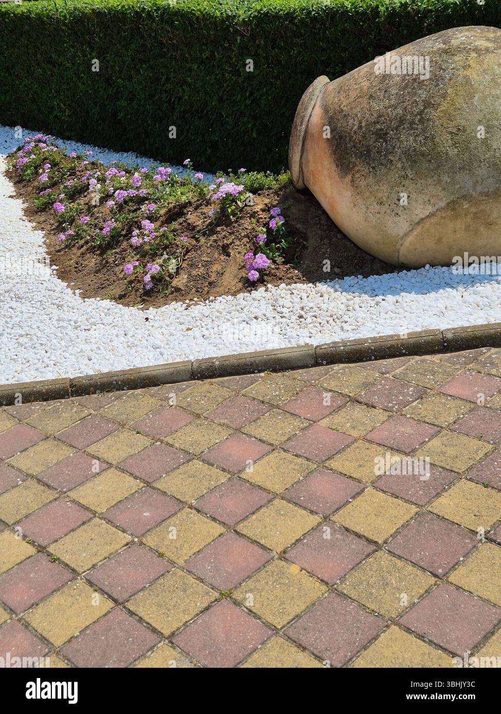 Eine antike Amphore, die einen Fluss aus zarten Blumen auf einen gepflasterten Weg „gießt“. Eine bezaubernde Gartenkomposition. Eine originelle Konstruktionslösung - Smartphone-aufgenommenes Stockfoto