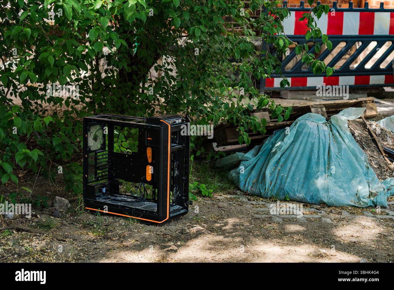 Ein schwarzes Computergehäuse mit orangefarbenen Akzenten befindet sich neben einer Baustelle. Üppiges Grün umgibt den weggeworfenen Gegenstand und eine blaue Plane Stockfoto Ein schwarzes Computergehäuse mit orangefarbenen Akzenten befindet sich neben einer Baustelle. Üppiges Grün umgibt den weggeworfenen Gegenstand und eine blaue Plane Stockfoto