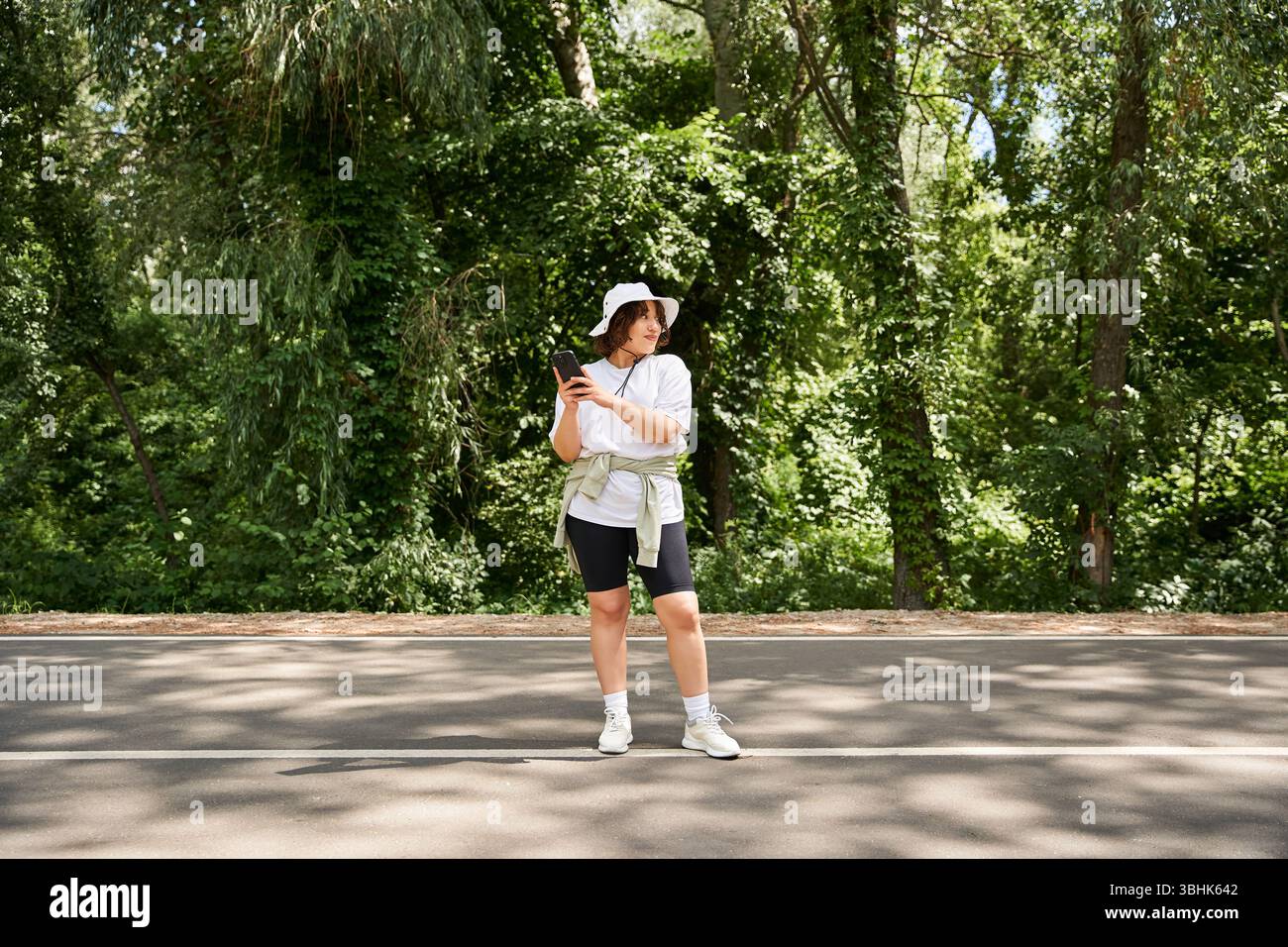Hell gekleidete Frau steht auf einem Pfad im Wald und genießt einen sonnigen Sommertag im Freien. Stockfoto