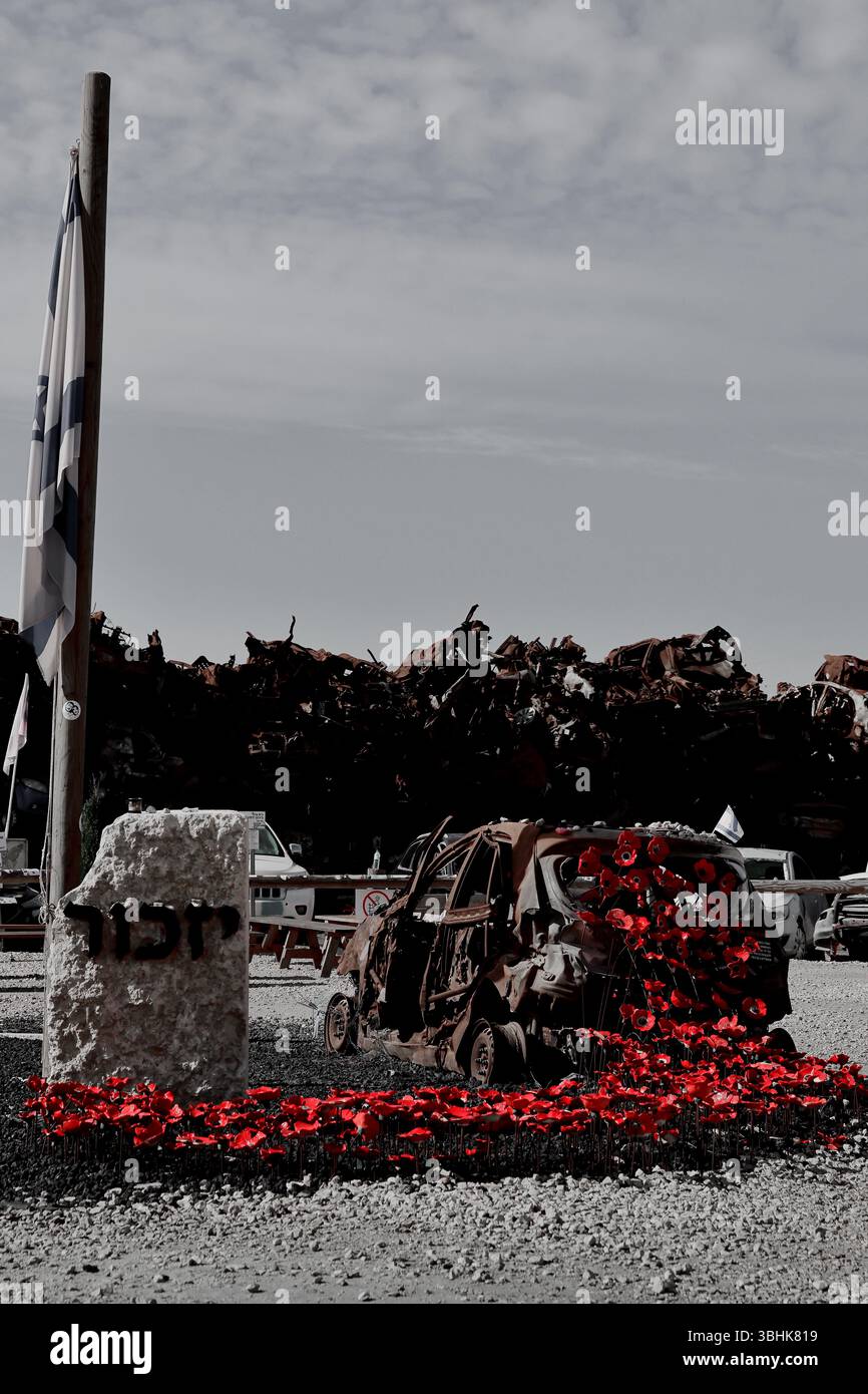 Ein verbranntes Fahrzeug mit rotem Keramikmohn steht hinter einem „Yizkor“-Stein an der Stelle der Car Wall vor einer feierlichen Mauer aus rostiger Zerstörung. Stockfoto