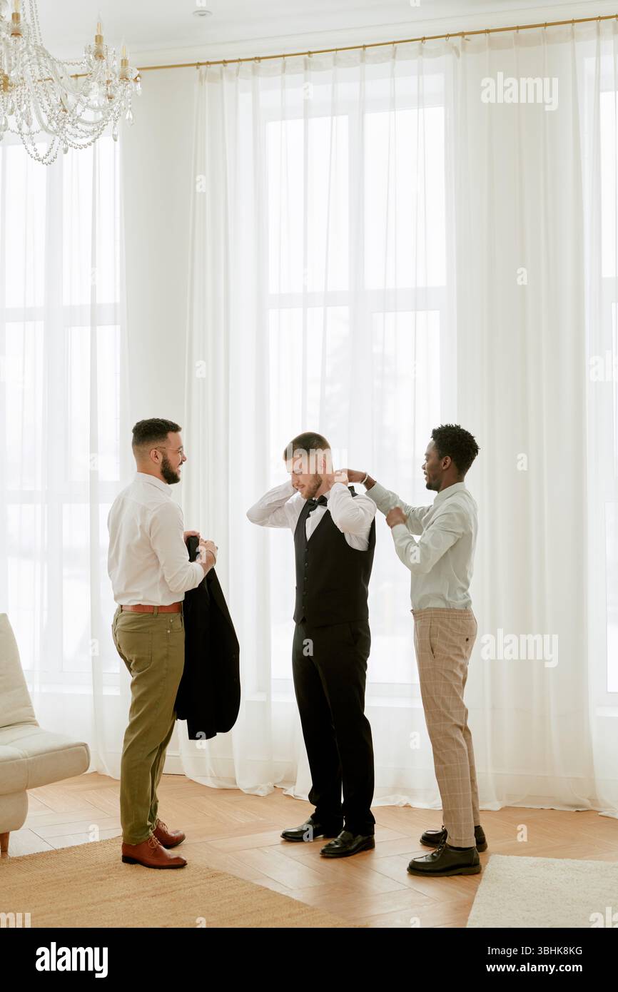 Drei junge Erwachsene Männer, zwei Kaukasier und ein Schwarzer, bereiten sich auf die Hochzeitsfeier vor, stehen zusammen in einem hellen Raum, ein Mann stellt eine Fliege ein, der dritte hält eine Jacke Stockfoto