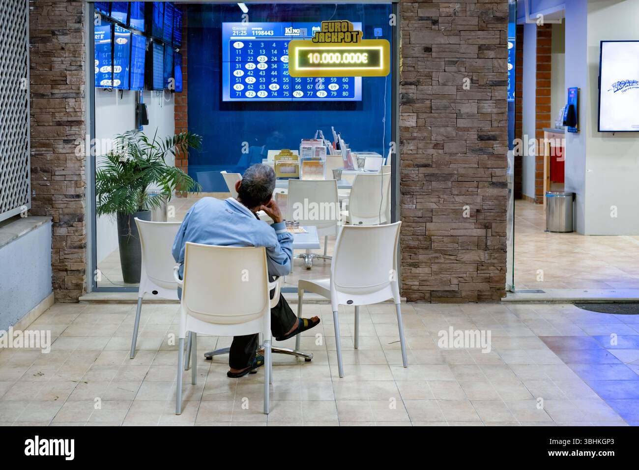 Rhodos Griechenland. Ein einsamer männlicher Spieler an einem Tisch vor einem Wettgeschäft. Auf dem Display des Shops ist ein Schild für die Wochen zu sehen, in denen die EuroMillions-Lotterie den Jackpot zieht Stockfoto