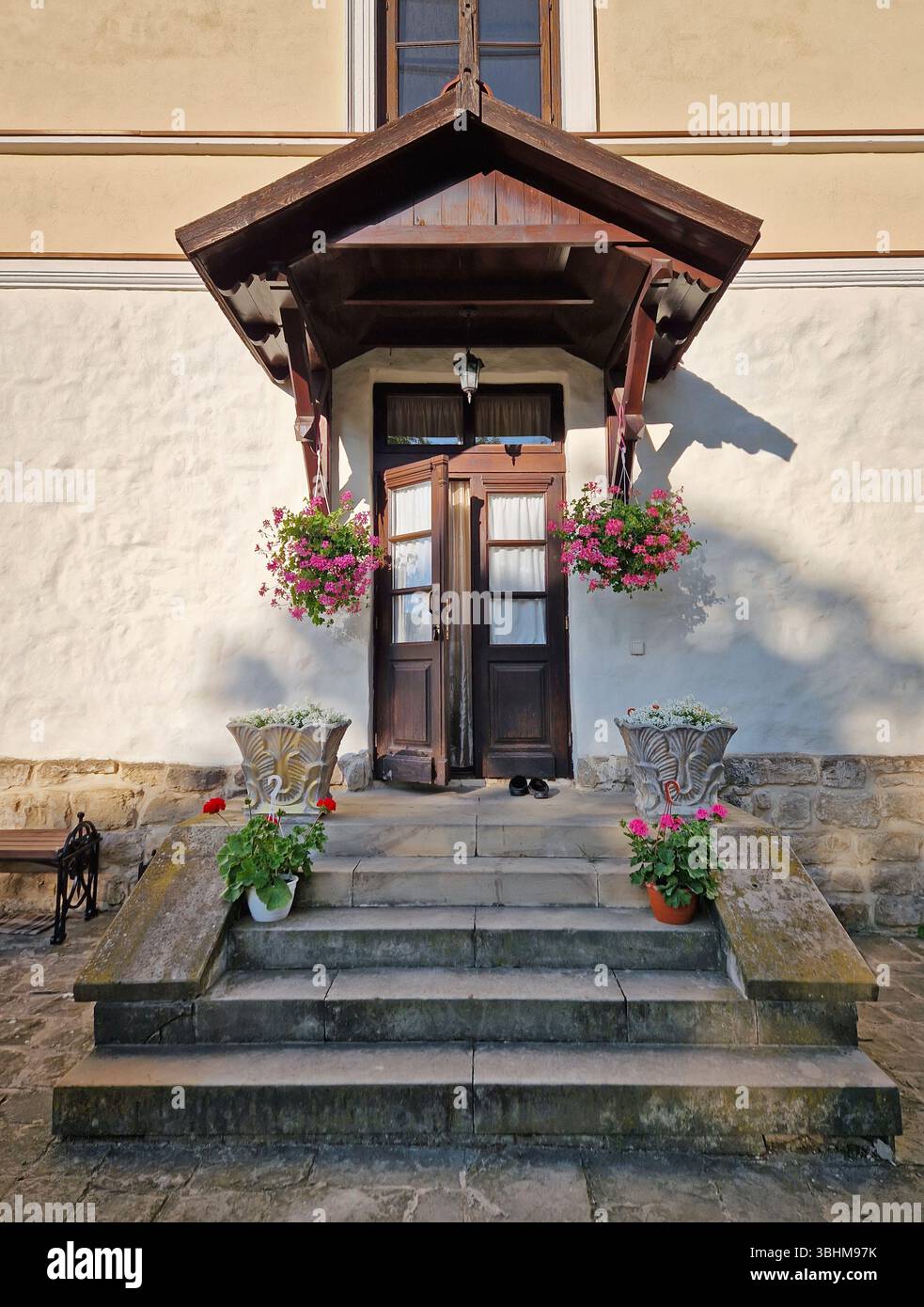 Rustikale alte Gebäudefassade mit Veranda und einer charmanten Holztür, eingerahmt von zwei Seitenfenstern und einem kleinen Überhang. Steintreppen führen zum Eingang - Smartphone-aufgenommenes Stockfoto