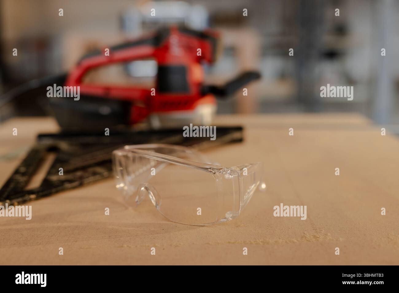 Klare Schutzbrillen liegen auf einer Holzwerkbank mit schwarzem quadratischem, rotem Schleifgerät auf Stockfoto