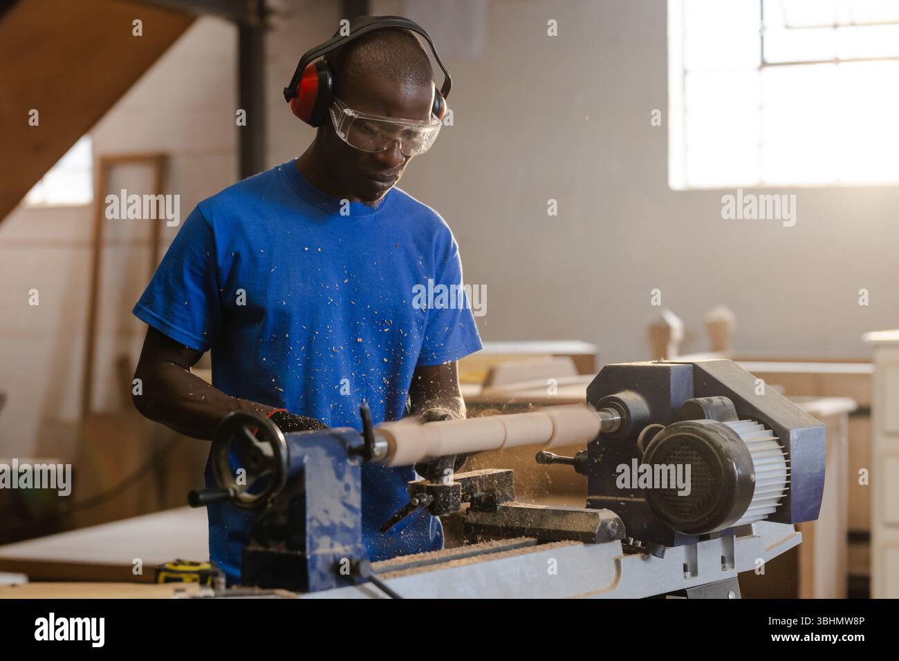 Afroamerikanischer Mann mit Gehörschutz und Schutzbrille, der Holz mit Meißel auf Drehmaschine in der Werkstatt rasiert Stockfoto