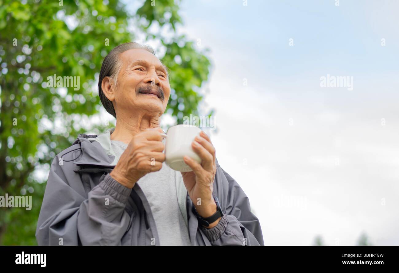 Hübscher älterer Mann trinkt, hält Kaffee aus einer großen Tasse, während er an einem Sommertag draußen am großen Baum steht. Kopierbereich. Stockfoto