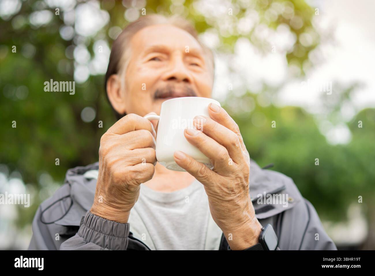 Gesunder asiatischer Seniorenmann trinkt Kaffee im Morgenpark. Selektiver Fokus auf die Hände. Getting Cup of Coffee Konzept. Stockfoto