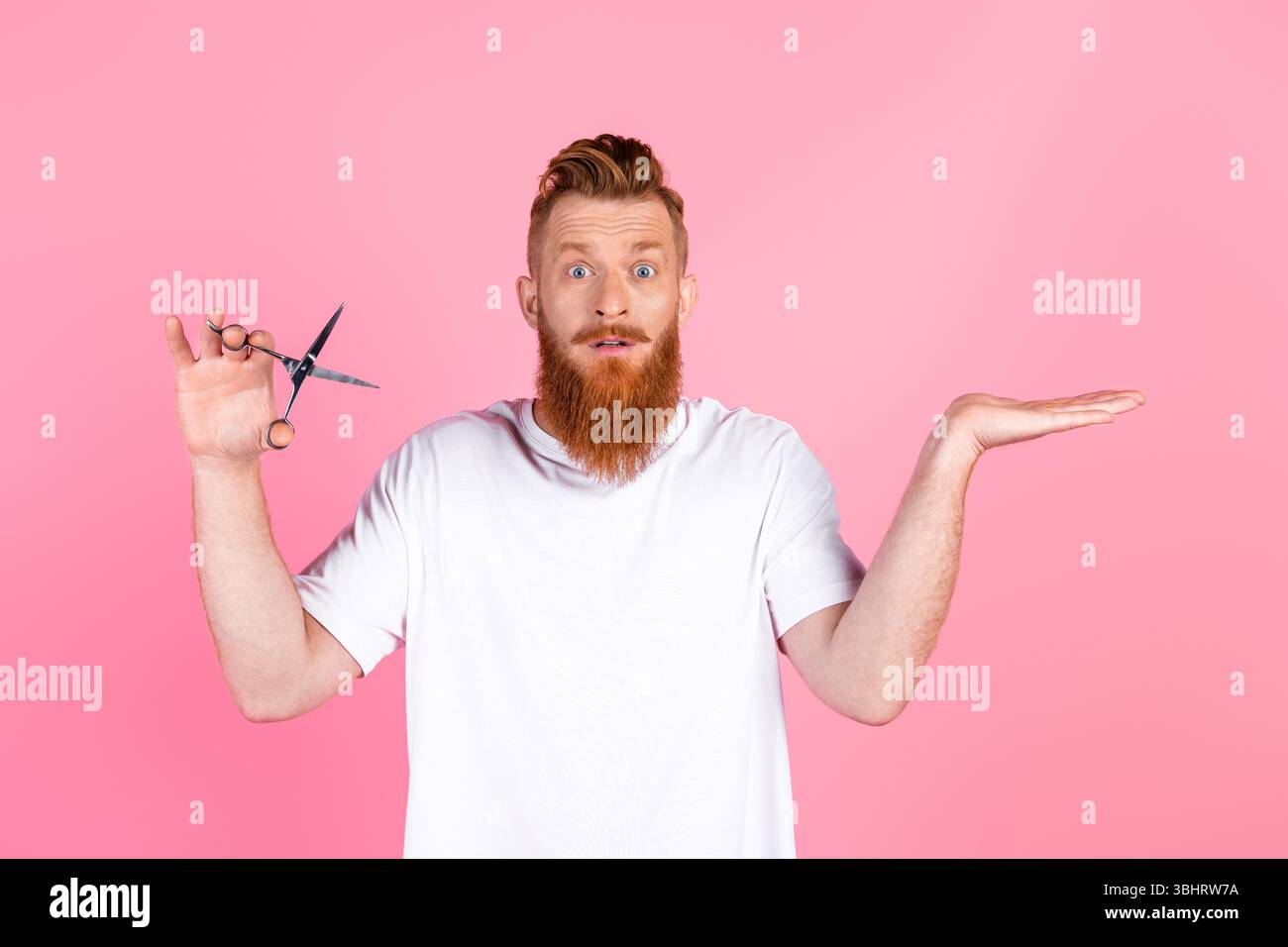 Fröhlicher bärtiger Mann in lässigem weißem T-Shirt mit Schere, Gesten auf rosa Hintergrund, verspielter Ausdruck Stockfoto