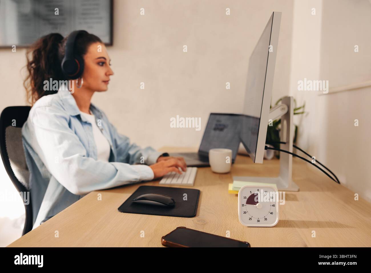 Eine junge Frau, die sich intensiv auf ihre Computerarbeit konzentriert, Kopfhörer eingeschaltet und einen Timer auf dem Schreibtisch sichtbar macht, um Produktivität und Konzentration zu veranschaulichen Stockfoto