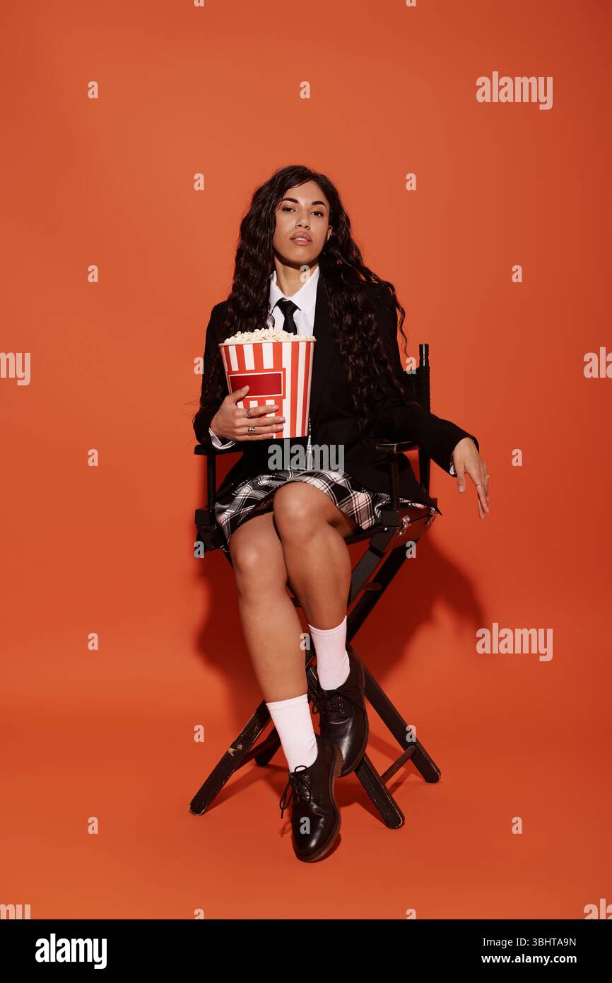 Eine junge Frau sitzt auf einem Stuhl mit Popcorn mit orangefarbenem Hintergrund und Schulmädchen-Kleidung. Stockfoto