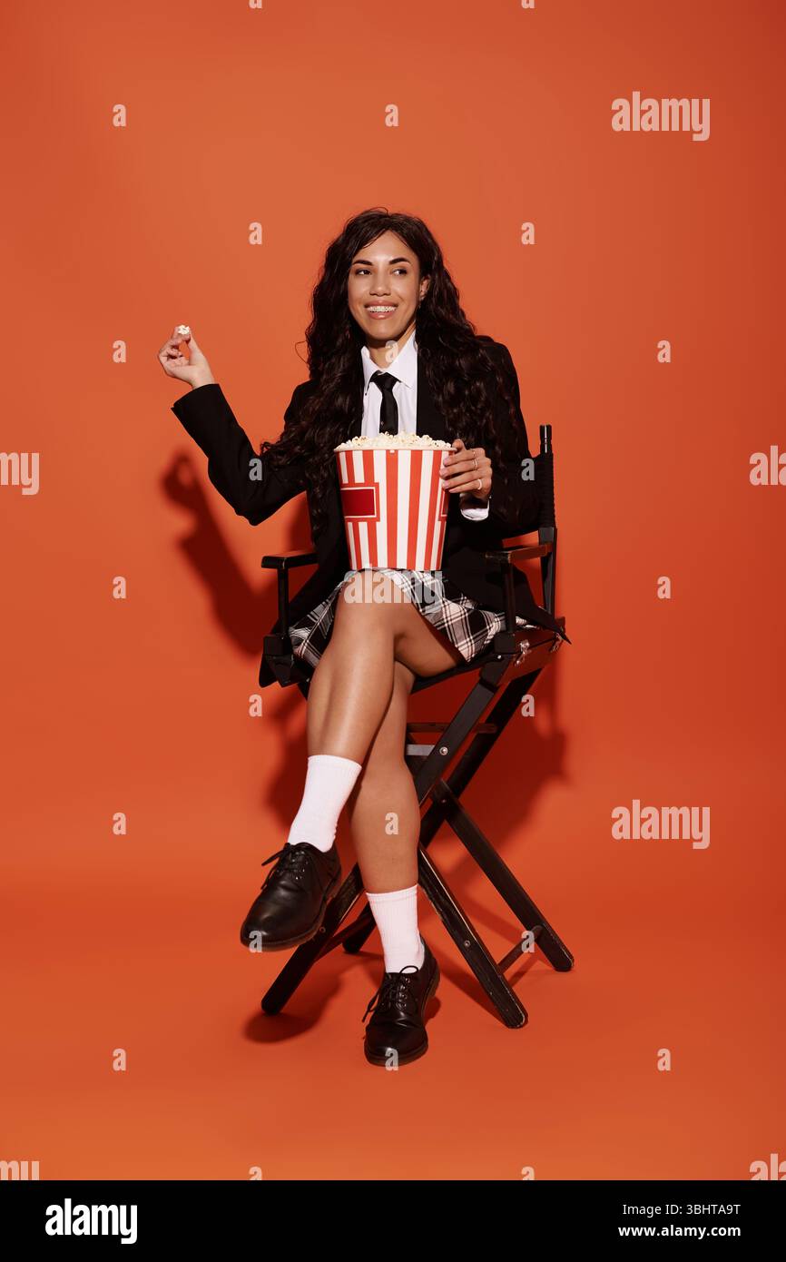 Eine junge Frau in Studentenkleidung sitzt mit Popcorn vor orangefarbenem Hintergrund. Stockfoto