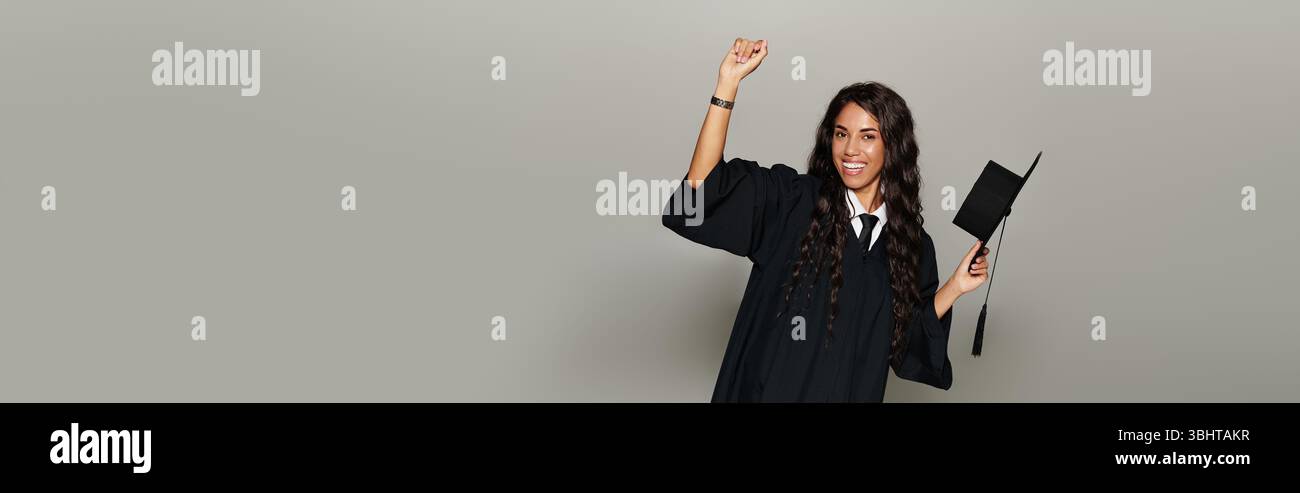 Eine fröhliche junge Frau in Abschlussmütze und Kleid feiert mit erhobenen Armen. Stockfoto