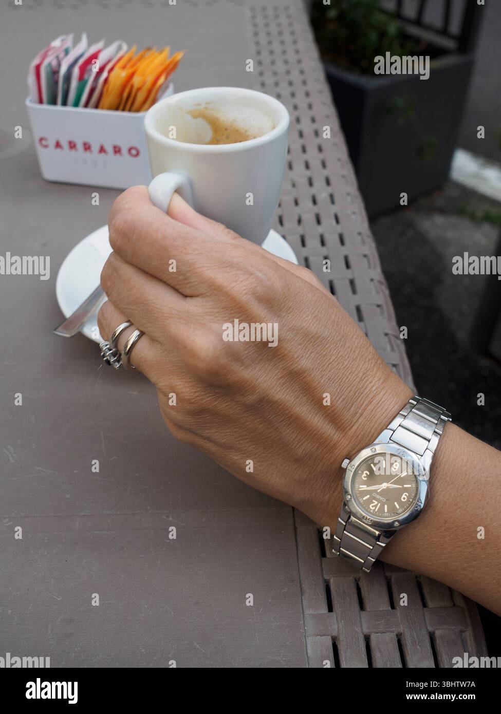Cremona, Italien 11. Juni 2025 Frau, die eine Armbanduhr trägt und eine Tasse Kaffee an einem Café-Tisch im Freien hält, mit Zuckerpackungen in der Nähe Stockfoto