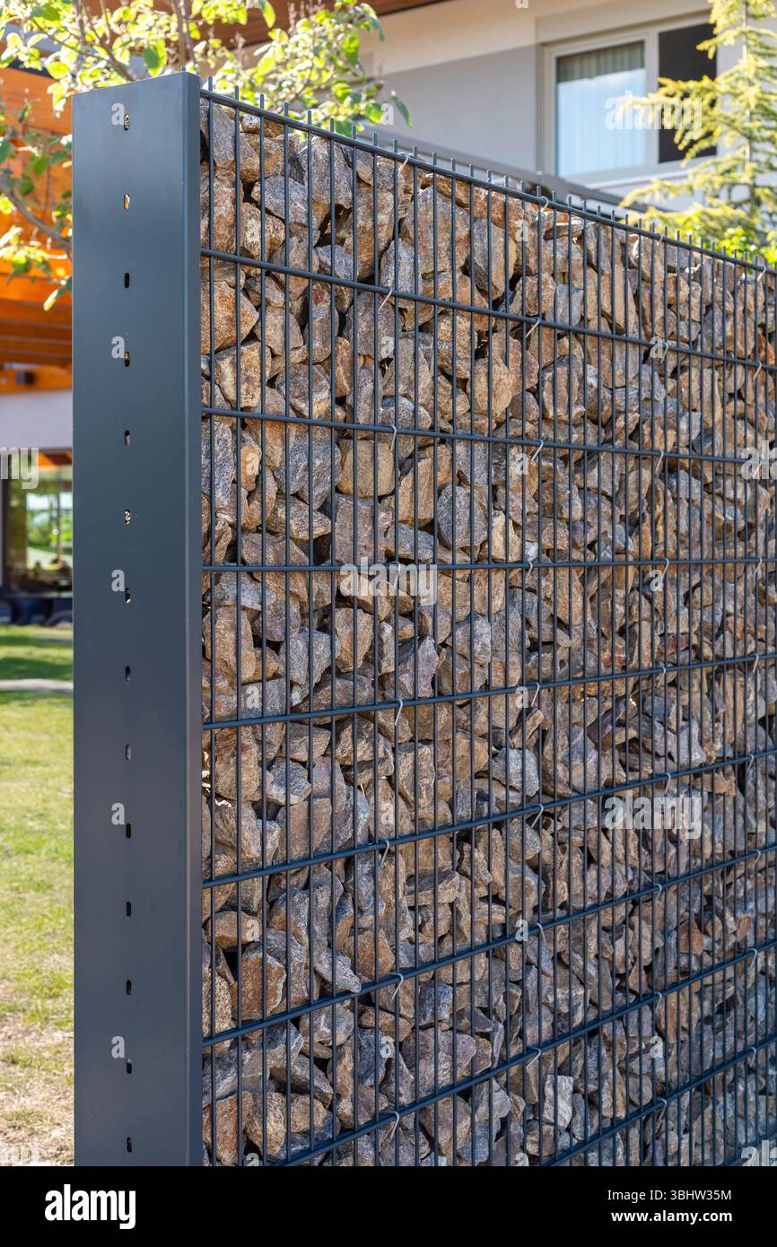 Ein einzigartiger Gabionenzaun aus Metalldrahtkäfigen mit Steinen steht in einem gut gepflegten Hof. Dieses moderne Design sorgt für eine besondere Note Stockfoto