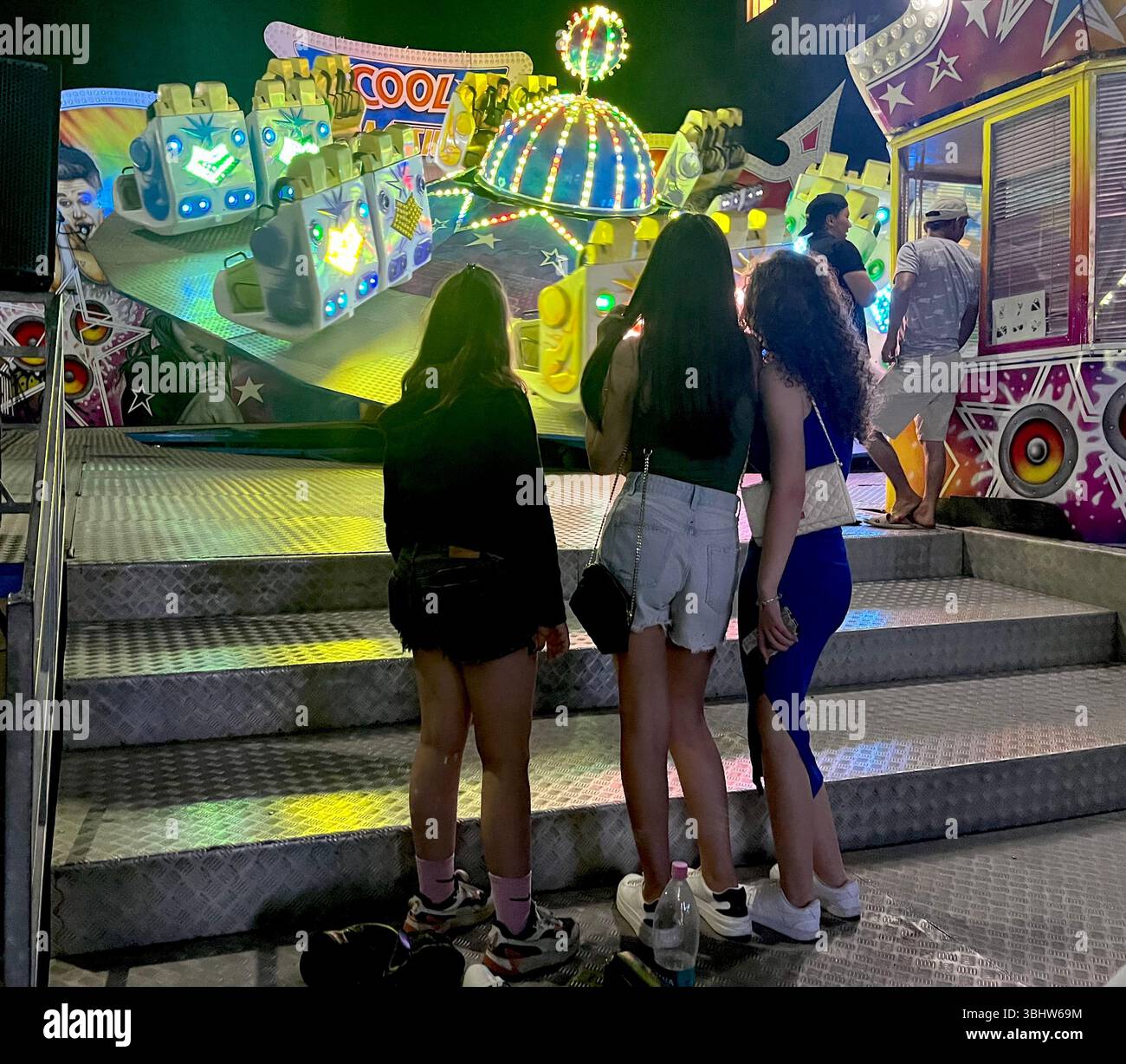 Mädchen genießen eine Nacht in einem Vergnügungspark vor einer hell beleuchteten Achterbahn, umgeben von bunten Lichtern und einer lebhaften Atmosphäre - Smartphone-aufgenommenes Stockfoto