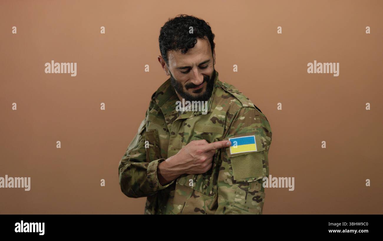 Hispanischer Mann in Militäruniform mit ukrainischem Flaggenpflaster, lächelt vor orangem Hintergrund. Stockfoto