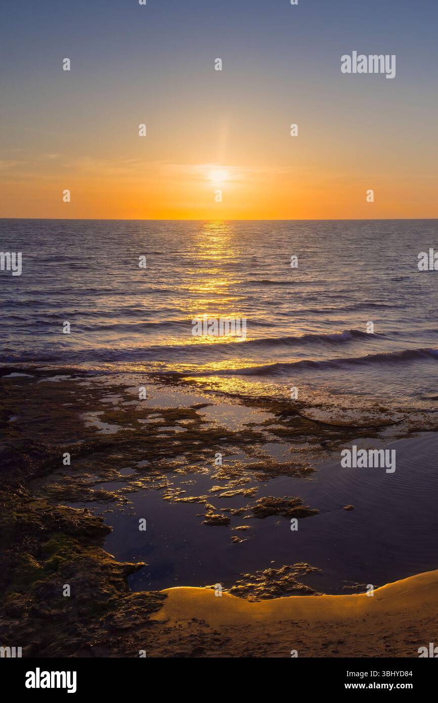 Goldenes Licht tanzt über dem Wasser, während Wellen sanft gegen eine ruhige Küste bei Sonnenuntergang schlagen. Stockfoto