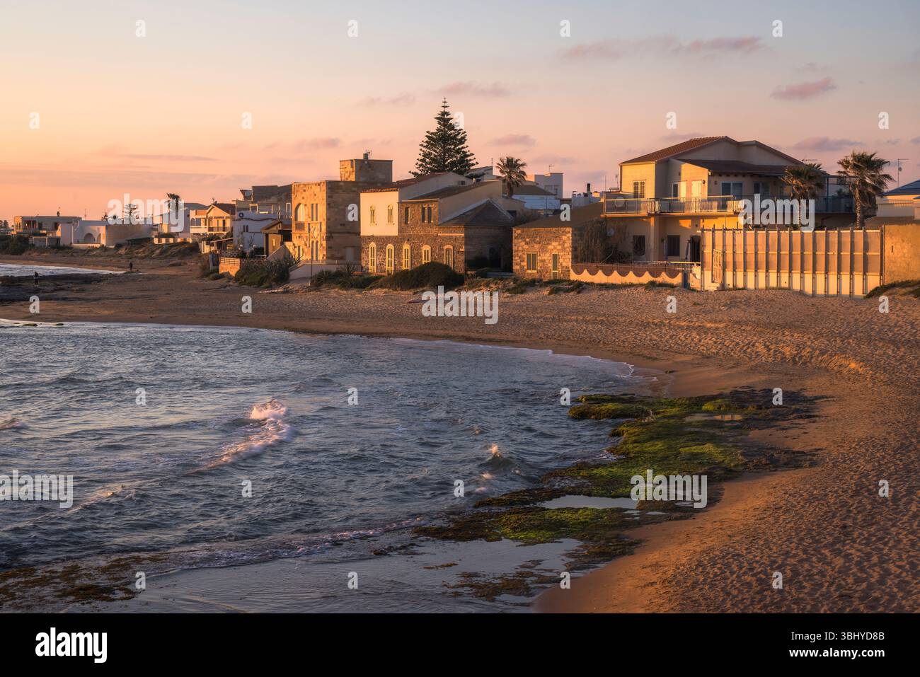 Goldenes Licht strahlt ein warmes Licht über den ruhigen Strand und unterstreicht den rustikalen Charme der Küstenhäuser von Punta Secca in Sizilien, Italien Stockfoto