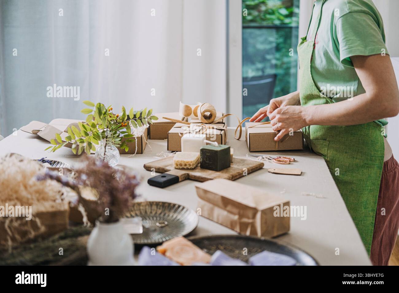Die Person bereitet Kraftseifenboxen in einem hellen Studio vor, das mit natürlichen Materialien und handgefertigten Produkten gefüllt ist. Langsam gemacht, achtsame Produktion, ethisch Stockfoto