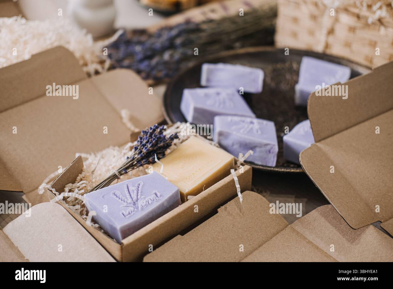 Lavendelseifen sind in offenen Kraftkästen mit geschreddertem Füllstoff auf einem Arbeitstisch angeordnet. Kunststofffreie Box, zerkleinerter Kraftfüller, nachhaltige Verpackung Stockfoto