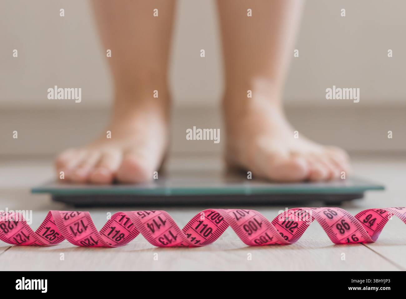 Ein Paar Füße steht auf einer digitalen Skala. Vor der Skala ist ein rosafarbenes Maßband aufgewickelt. Die Umgebung ist hell und minimalistisch, wobei der Schwerpunkt auf h liegt Stockfoto