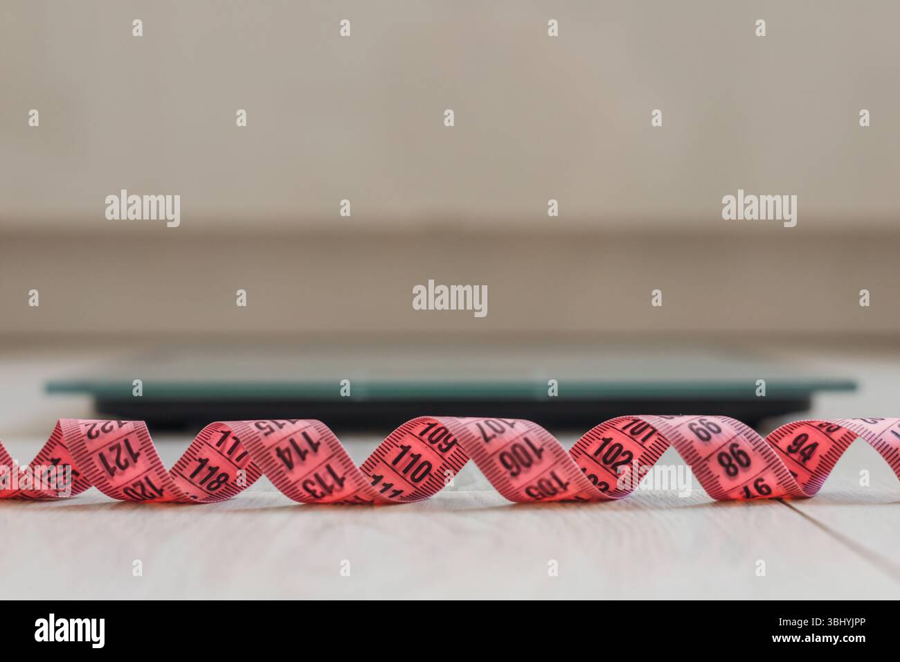 Eine Nahaufnahme eines rosafarbenen Maßbandes, das auf einem Holzboden aufgerollt ist, mit einer digitalen Skala im Hintergrund. Die Szene setzt auf Gesundheits- und Fitnesskonzepte Stockfoto