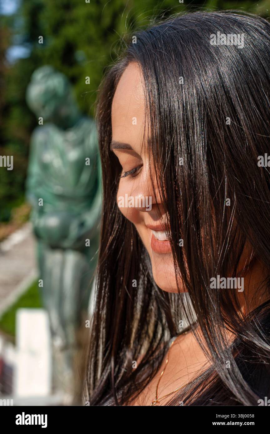 Nahporträt einer lächelnden jungen Frau mit langen dunklen Haaren, die teilweise ihr Gesicht bedecken. Sie genießt einen sonnigen Tag im Freien, mit grüner Bronze Stockfoto