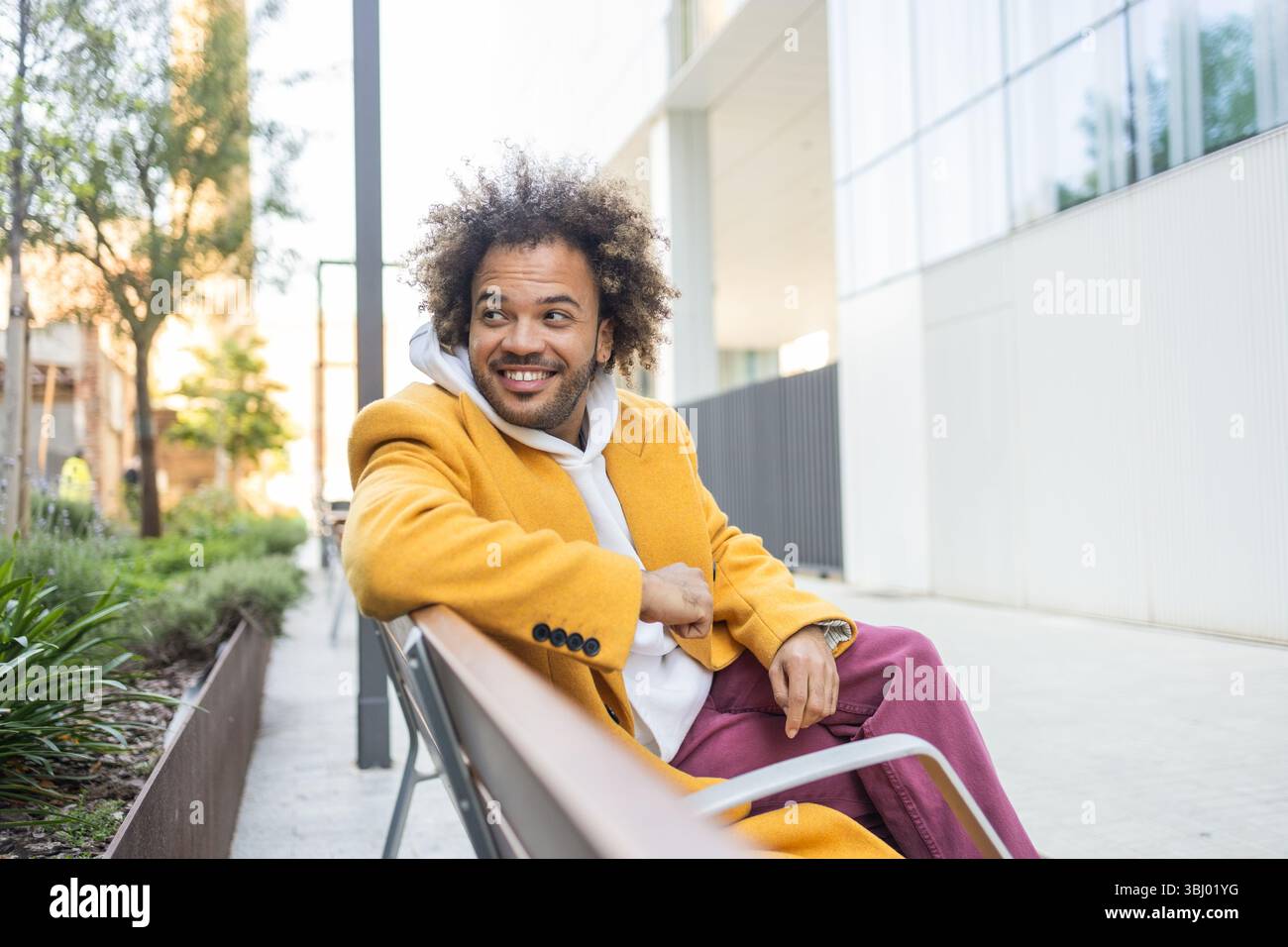 Ein fröhlicher Afro-Mann sitzt bequem auf einer Bank in einer modernen urbanen Umgebung, trägt einen stilvollen gelben Mantel und eine lila Hose und genießt einen Moment der Entspannung Stockfoto