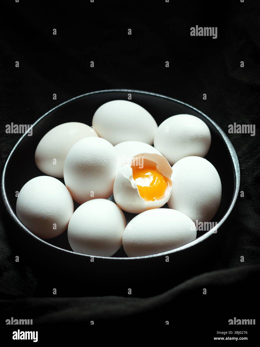 Eine Sammlung weißer Eier liegt in einer dunklen Schüssel auf schwarzem Hintergrund, wobei ein Ei aufgerissen wird, um sein hellgelbes Eigelb freizugeben, und ein P hinzufügt Stockfoto