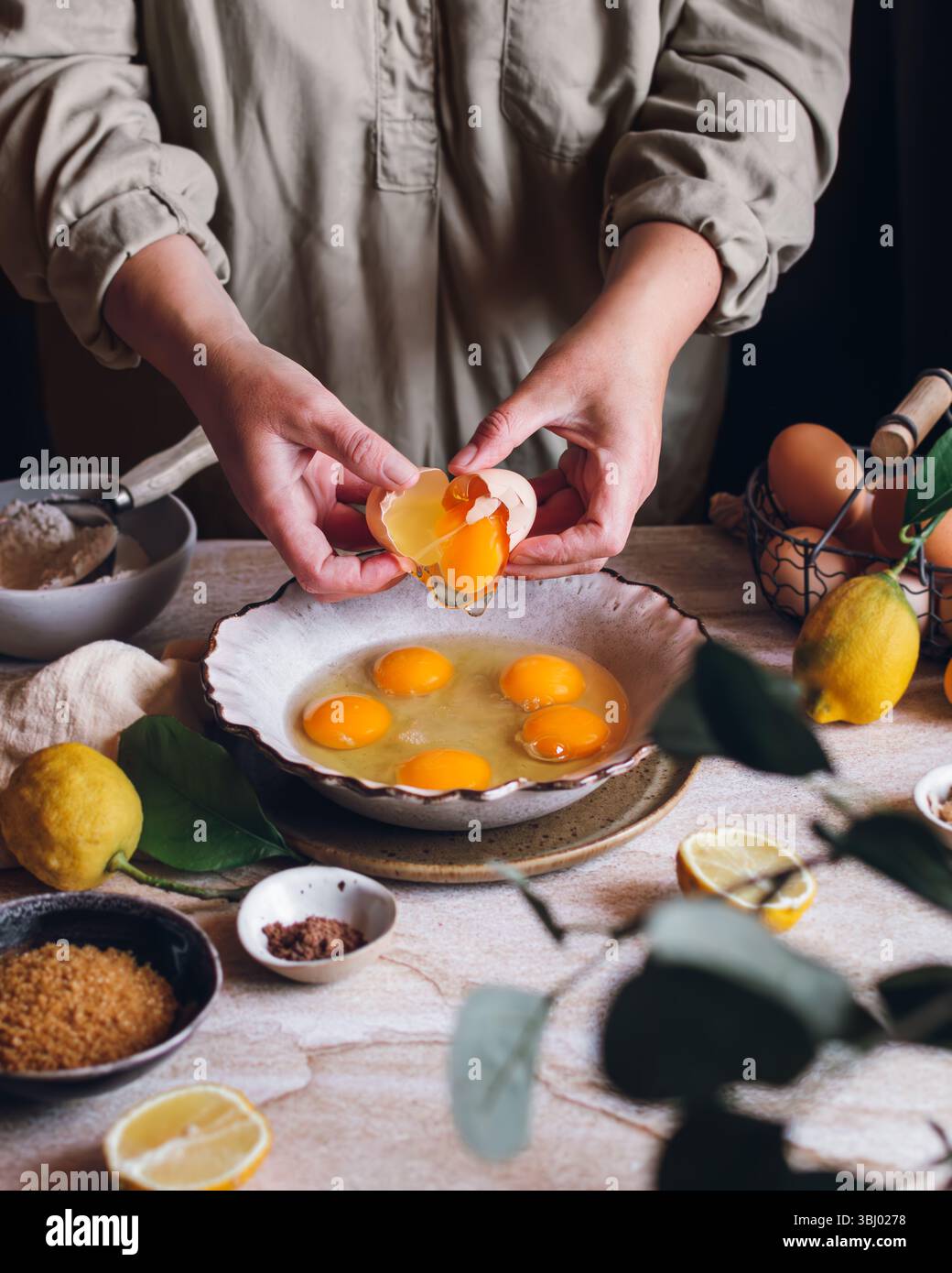 Eine Person knackt Eier in eine Schüssel auf einem Tisch, umgeben von Zitronen, braunem Zucker und Gewürzen, um eine rustikale Küchenszene voller frischer Zutaten einzufangen Stockfoto