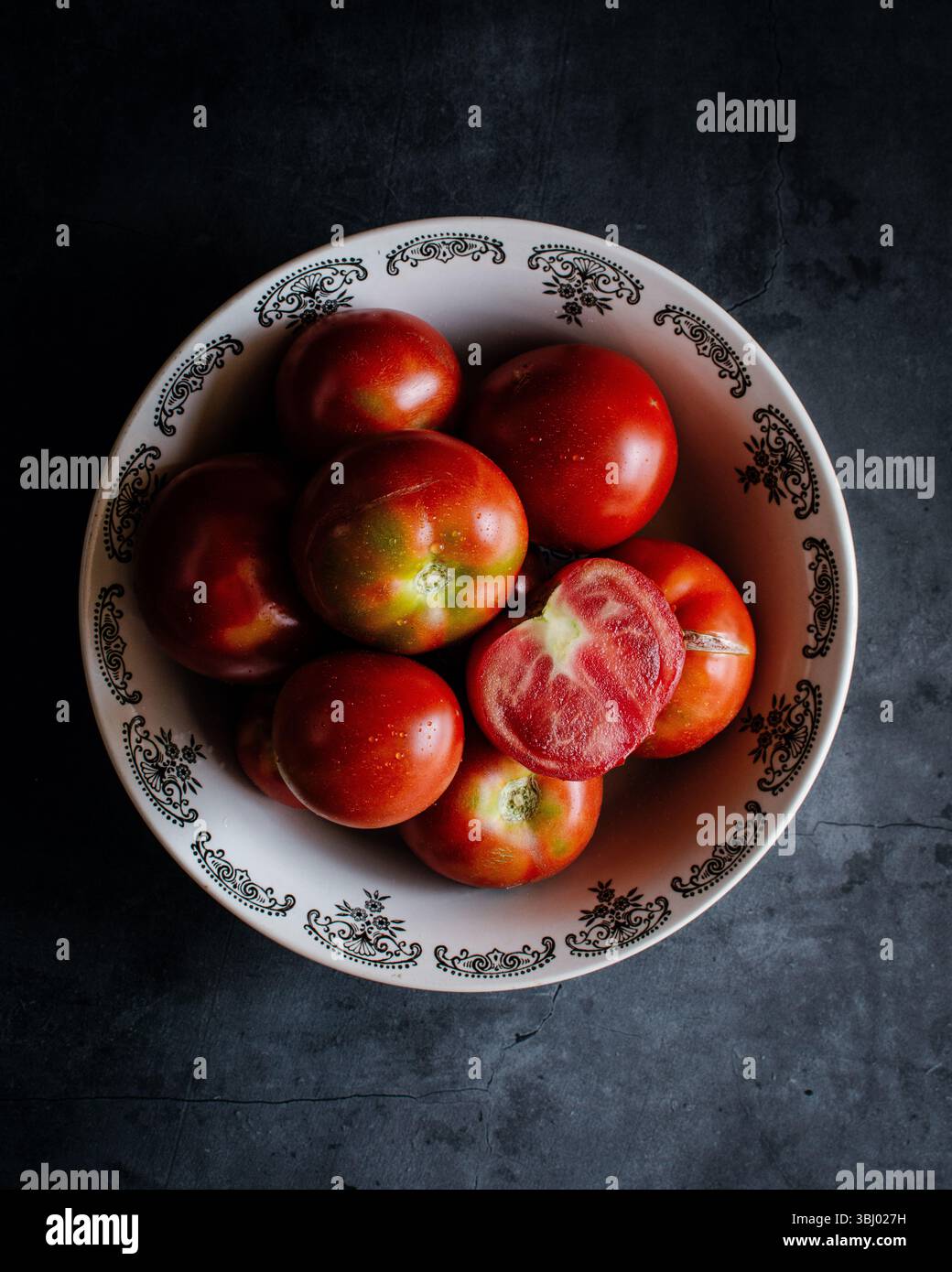 Ein Blick von oben nach unten auf frische, lebendige Tomaten in einer dekorativen Keramikschale vor dunklem Hintergrund. Eine Tomate wird in Scheiben geschnitten und zeigt ihre saftige Note Stockfoto
