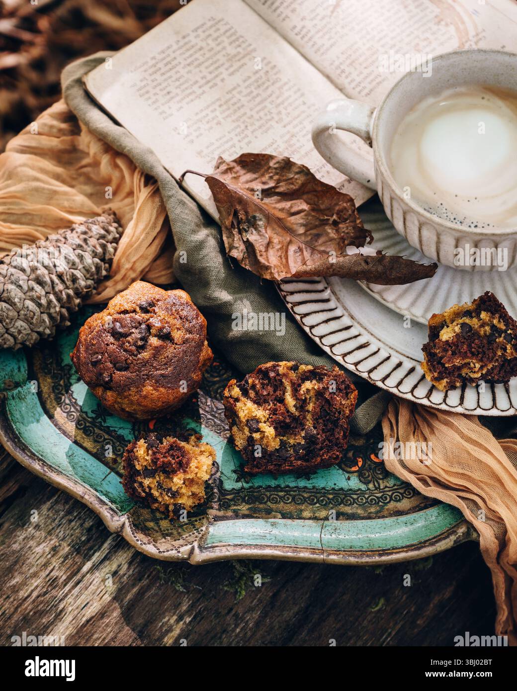 Eine gemütliche Herbstkulisse mit Kürbismuffins, einer warmen Tasse Kaffee und einem offenen Buch. Die Szene ist mit rustikalen Elementen wie Tannenzapfen bereichert Stockfoto
