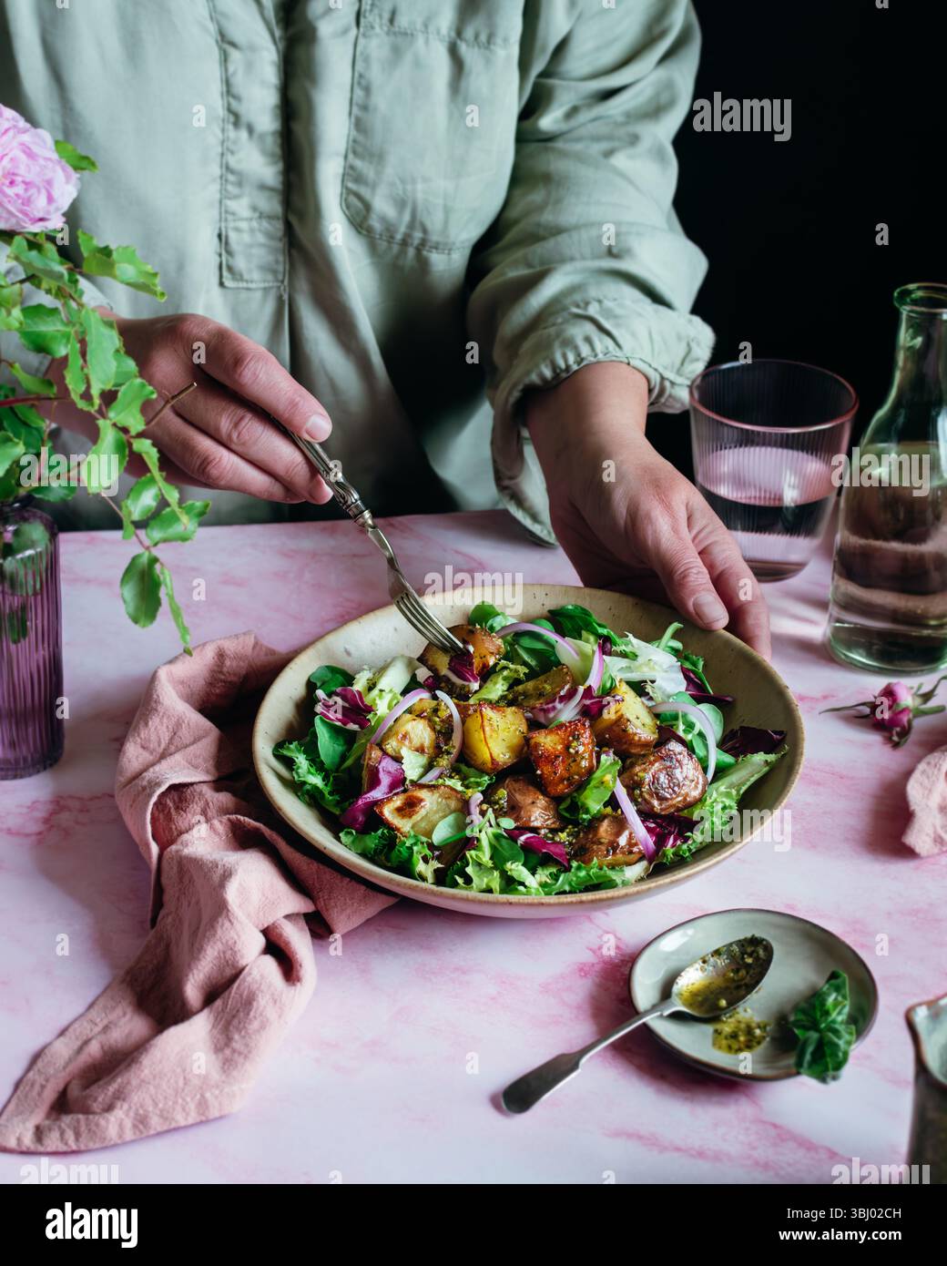Eine Person in einem hellen Hemd bereitet einen lebendigen Kartoffelsalat mit frischem Gemüse, gerösteten Kartoffeln und roten Zwiebeln auf einem rosafarbenen Tisch zu. Eine Gabel wird gehalten, bereit für Stockfoto