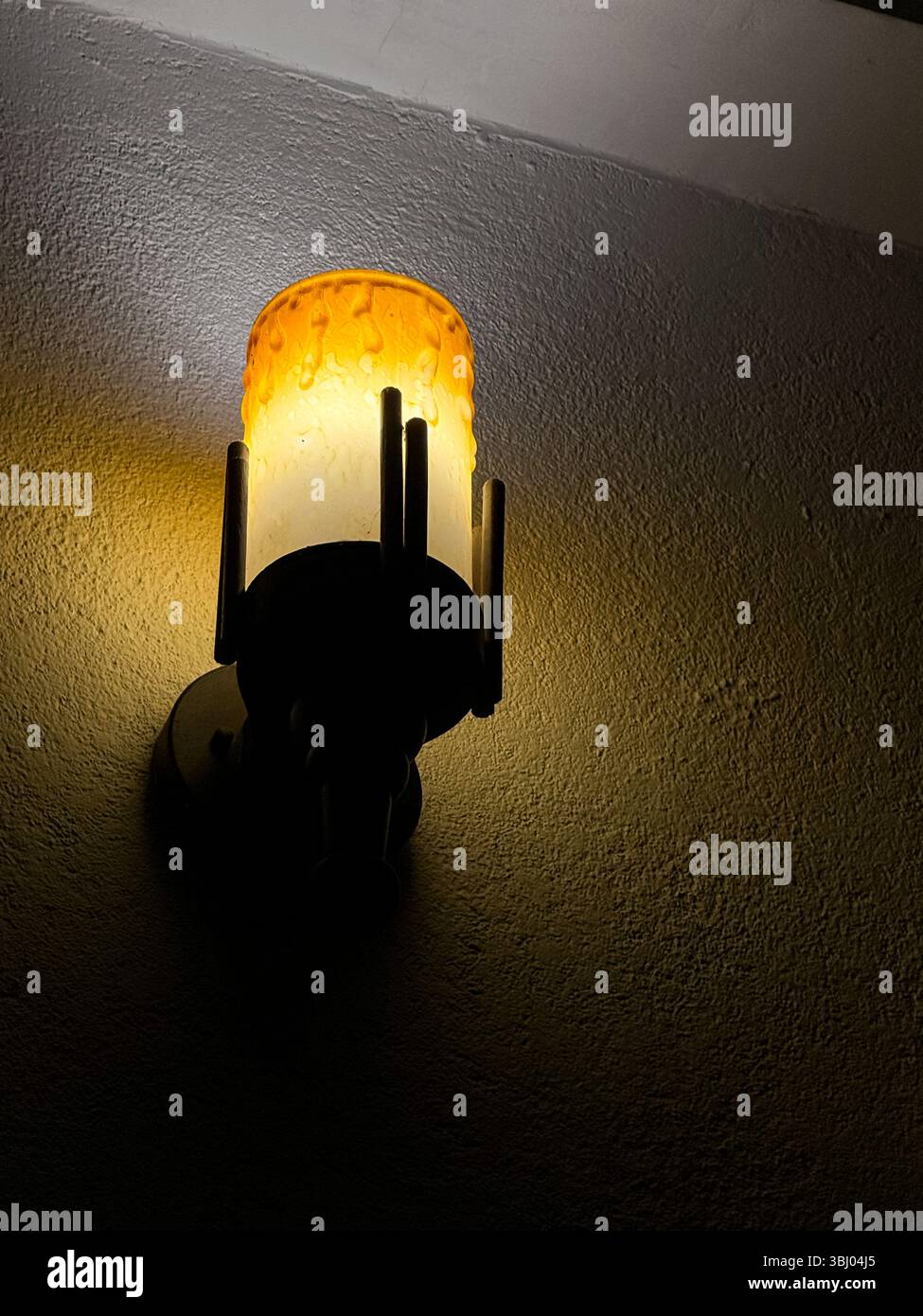 Eine leuchtende Wandleuchte mit einem strukturierten bernsteinfarbenen Glasschirm, die nachts warmes Licht auf eine raue Wand wirft. - Smartphone-aufgenommenes Stockfoto