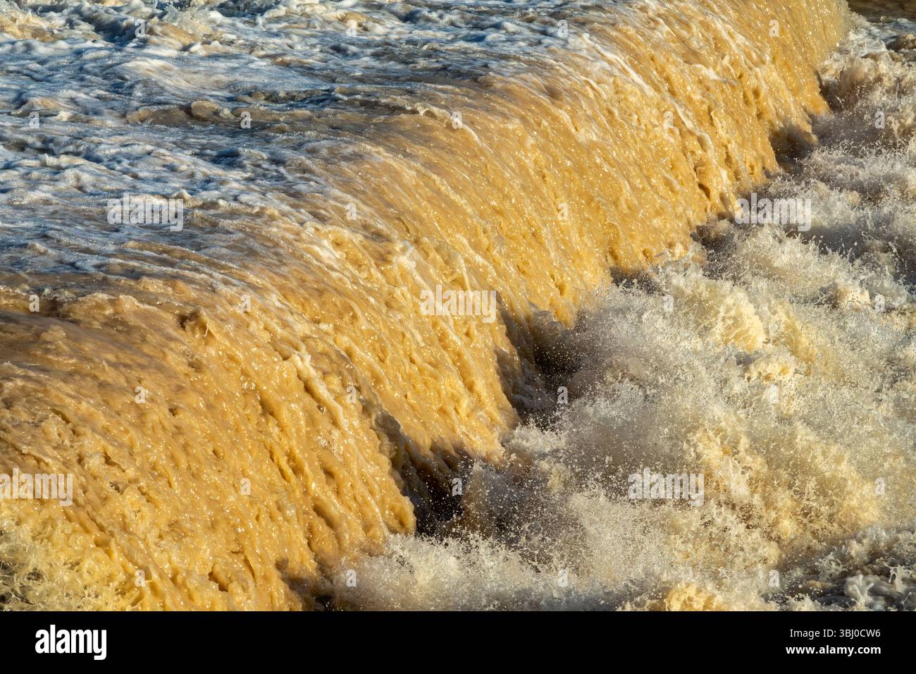 Dynamische Nahaufnahme eines kaskadierenden Wasserfalls, gefüllt mit Schlammwasser, aufgenommen während einer Hochwassersaison. Die starke Strömung und das dicke Sediment erzeugen Stockfoto