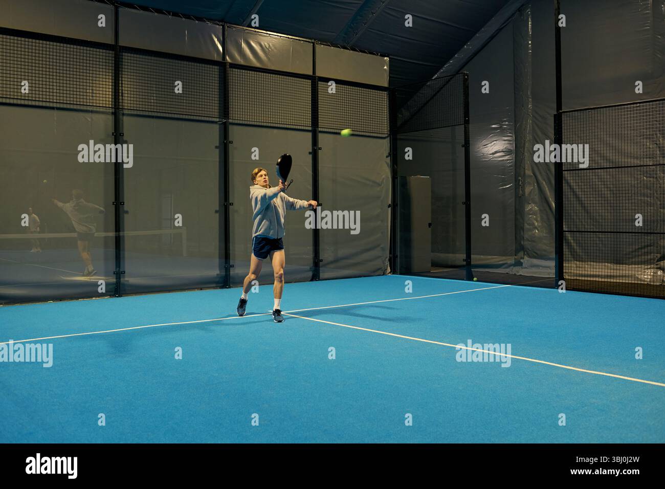Ein engagierter junger Mann trainiert mit Entschlossenheit auf einem Indoor-Padel-Platz und konzentriert sich auf seinen nächsten Schuss. Stockfoto
