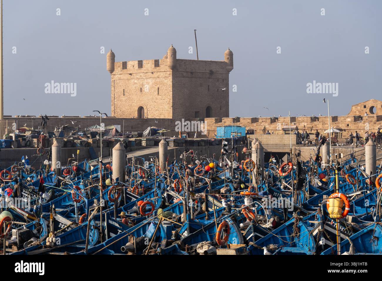 Essaouira, Marokko - 21. Februar 2025: Traditionelle Fischerboote liegen im Hafen von Essaouira mit dem Turm der Festung Mogador in Mor vor Anker Stockfoto