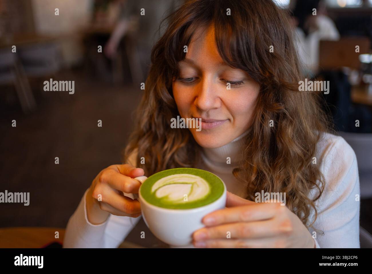 Junge Frau, die in einem gemütlichen Café einen warmen Matcha-Latte mit komplizierter Blattkunst genießt und die friedliche Atmosphäre und die reichen, erdigen Aromen des grünen Teegetränks genießt Stockfoto