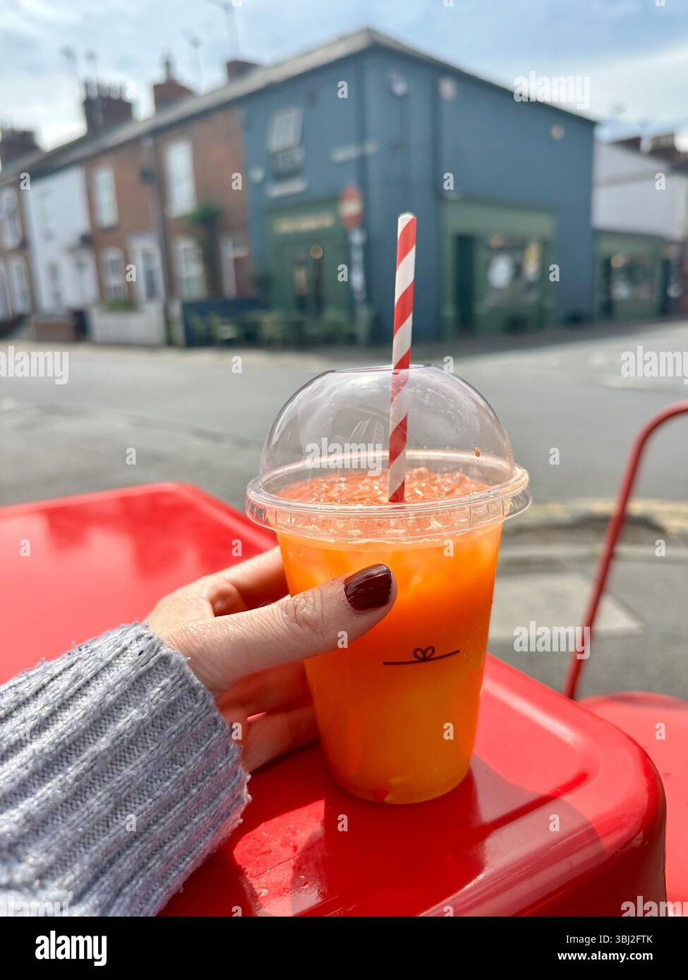 Karotten- und Ingwersaft zum Mitnehmen mit gestreiftem Strohhalm in der Hand mit dunkelrotem Nagel, beigefarbenem Cardiganärmel, rotem Tisch und Café im Hintergrund - Smartphone-aufgenommenes Stockfoto