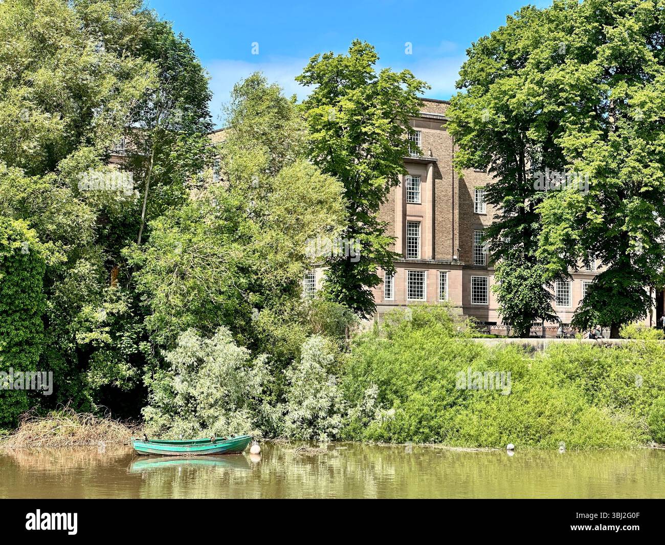 Leeres, grün lackiertes Ruderboot auf dem River Dee in Chester mit üppigen Bäumen und historischem Universitätsgebäude im Hintergrund an einem sonnigen Tag - Smartphone-aufgenommenes Stockfoto