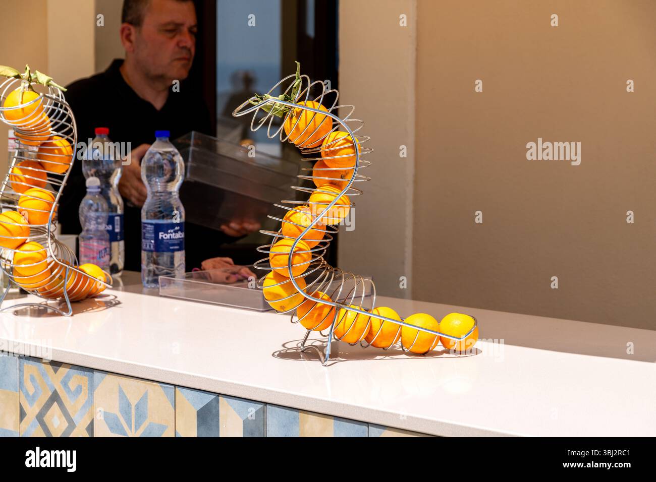 Taormina, Sizilien - Italien - 05-04-2025: Ausstellung mit Orangen in einem Metallspiralhalter an einem Theken in Taormina, Sizilien, mit einem Mann und Flaschen Wasser Stockfoto