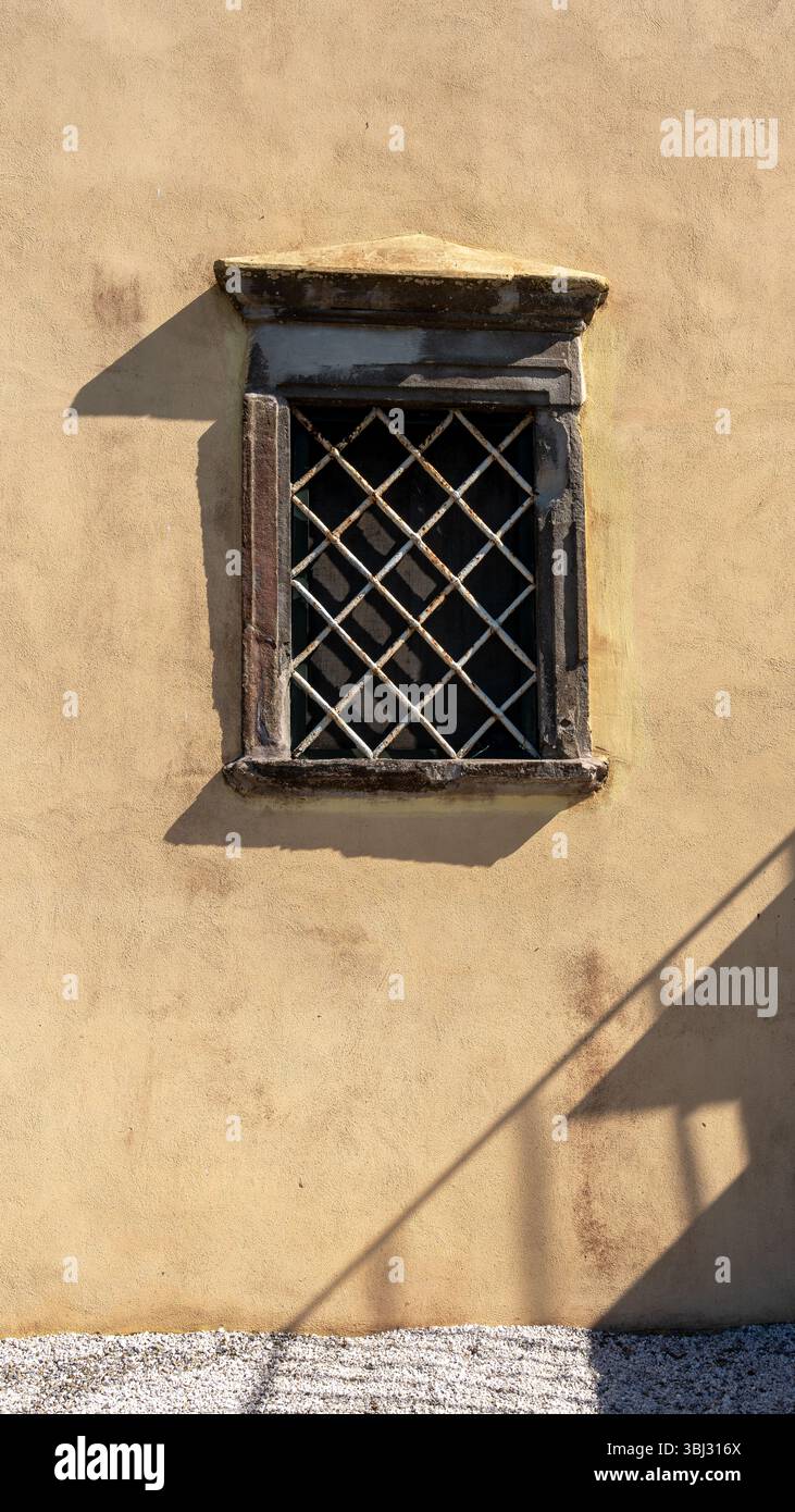 Altes Steinfenster mit Sicherheitsgitter an einer sonnendurchfluteten Stuckwand Stockfoto