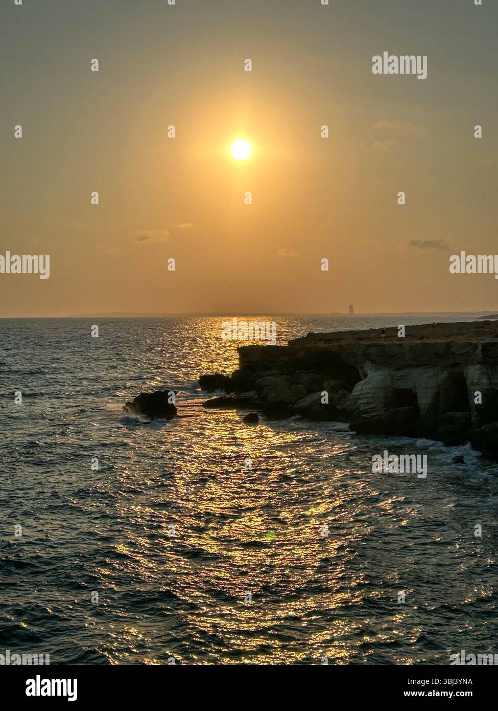 Goldene Sonne untergeht über dem Meer, wirft warme Reflexe auf das Wasser und beleuchtet die zerklüfteten Küstenklippen. Eine ruhige und dramatische Aussicht auf das Meer. - Smartphone-aufgenommenes Stockfoto