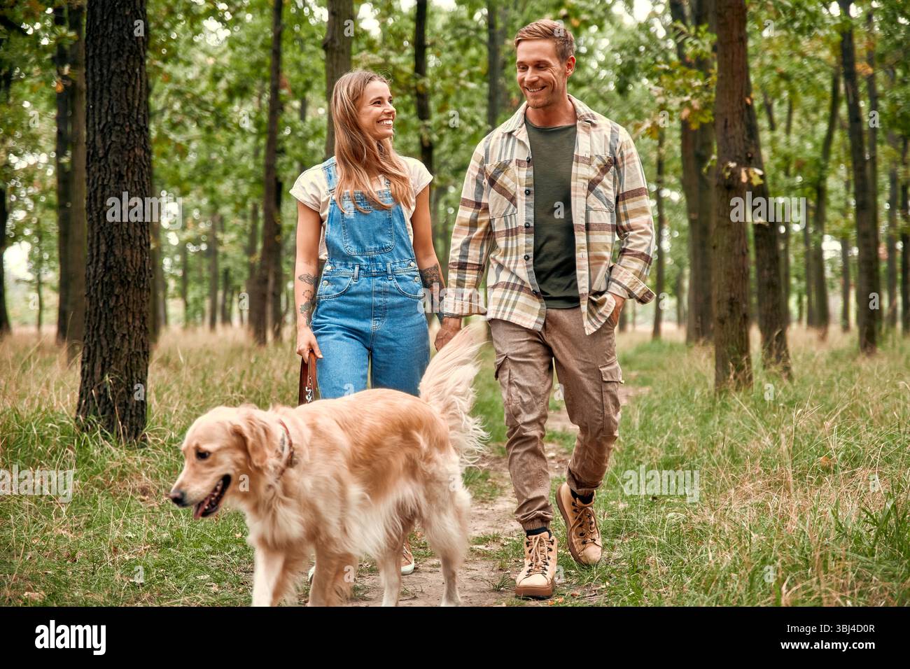 Ein fröhliches Paar schlendert Hand in Hand durch einen wunderschönen üppigen Wald, glücklich begleitet von ihrem charmanten Golden Retriever, während sie sich an dem erfreuen Stockfoto